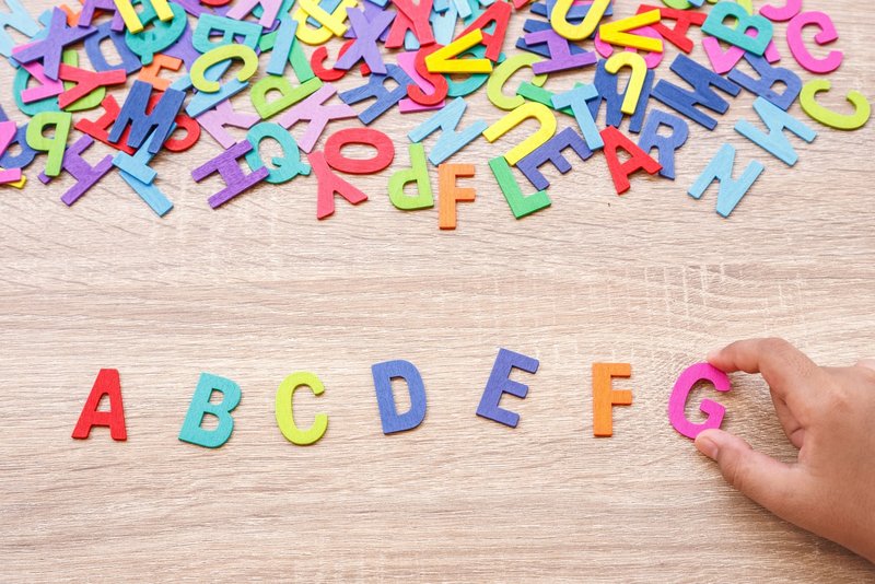 Bunte Buchstaben auf Tisch, Hand setzt den Buchstaben G in die Reihe A–F