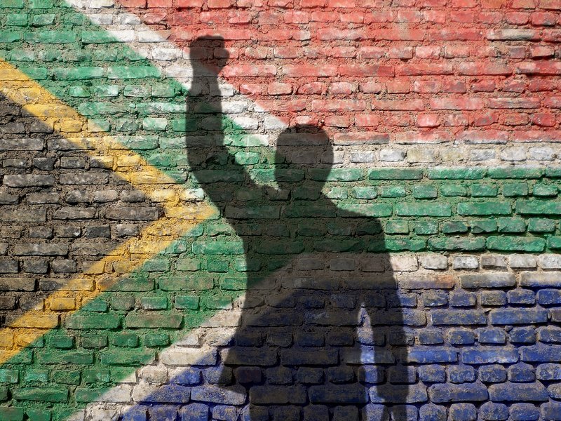 Der Schatten einer Person die ihre Faust hochhebt, vor der Flagge von Südafrika.